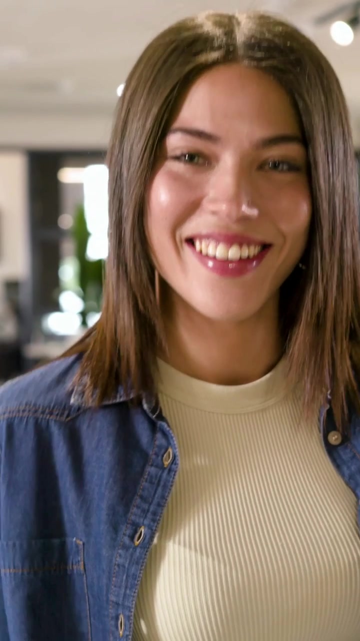 Video vertical: Mujer sonriendo, dando pasos, inclinándose y recogiéndose el cabello mientras presenta en la oficina, vestida con mezclilla.