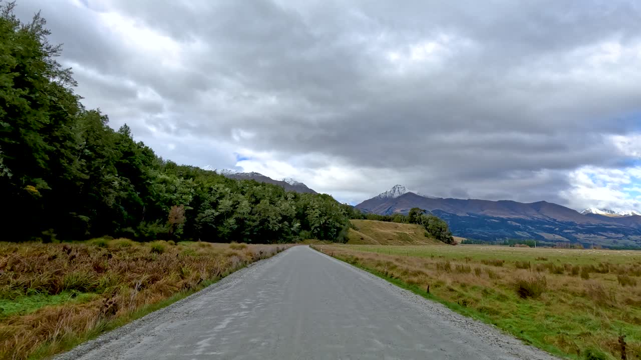 Vehicle drives along rural gravel road past forest and fields under overcast sky, steady motion
