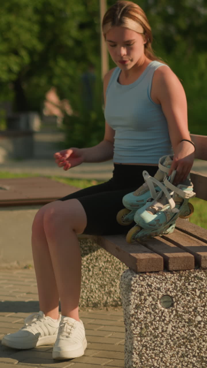 젊은 여성이 야외의 나무 벤치에 시안 롤러 스케이트를 놓고, 그녀는 검은색 쇼트와 색 스니커를 입고 있으며, 배경에는 나무, 초록색, 밝은 빛 아래의 여러 벤치가 있습니다.