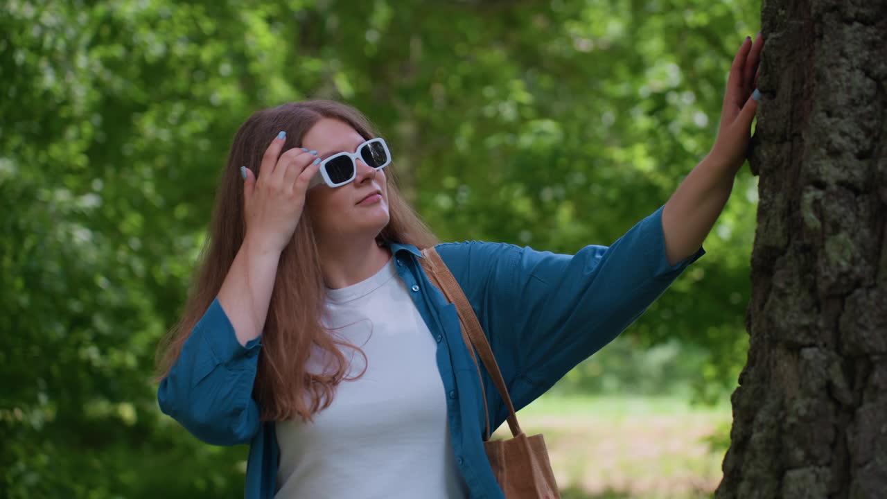 Una joven camina hacia el tronco de un árbol alto y toca su corteza mientras observa de cerca la textura, rodeada de un exuberante follaje verde bajo la luz del sol, sintiendo una suave brisa y paz.
