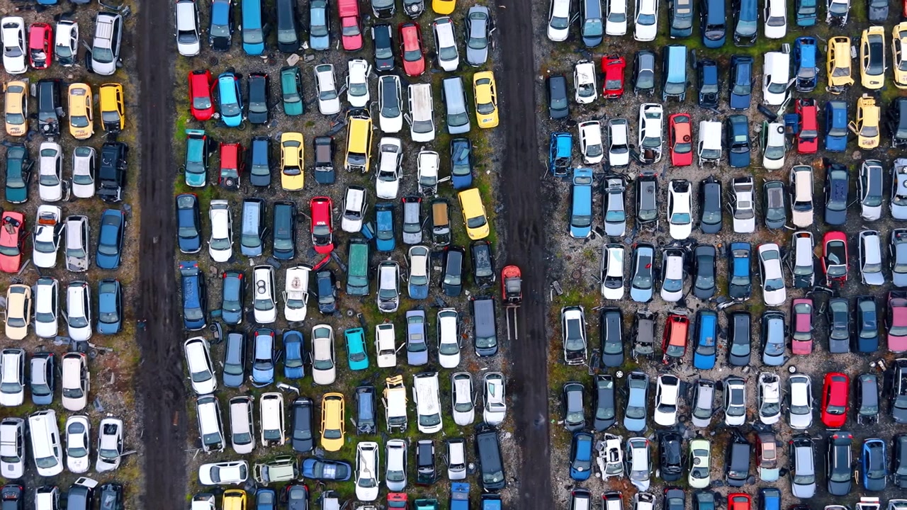 Prospettiva aerea verticale di un deposito di riciclaggio veicoli. Inquadratura simmetrica dall'alto di auto colorate parcheggiate in colonne verticali con un piccolo carrello elevatore in funzione.