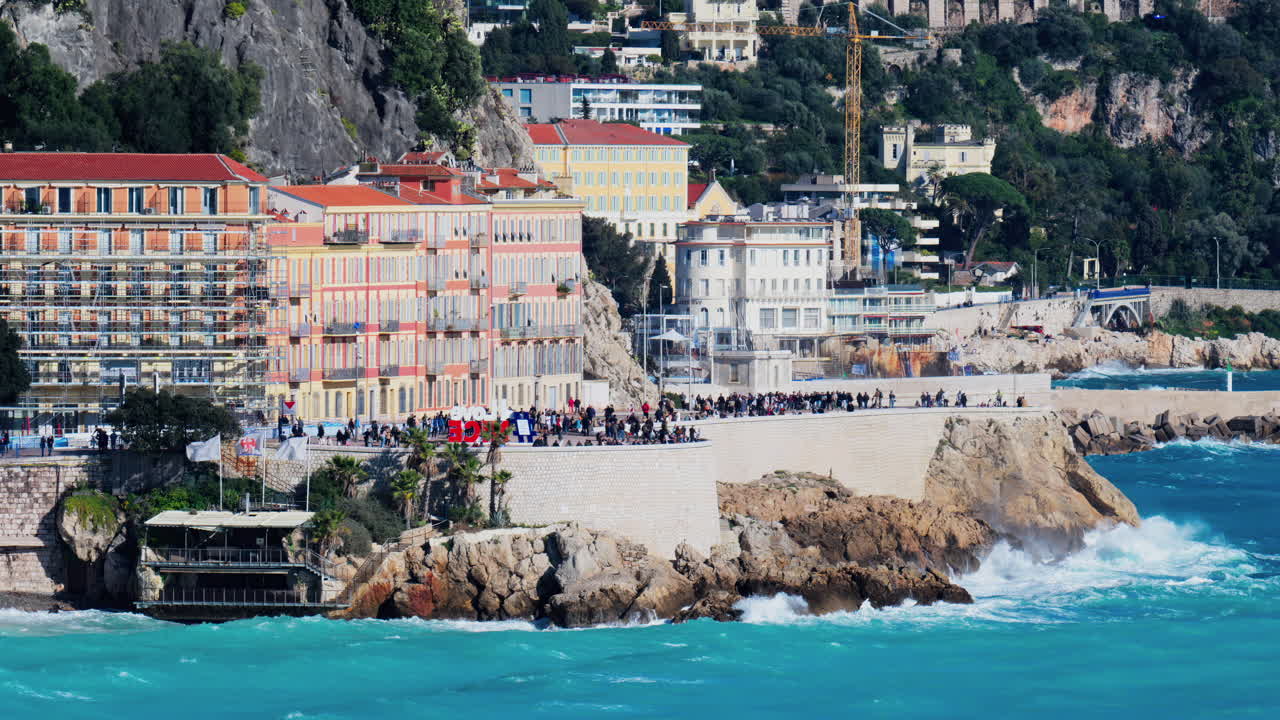 Nice, France - March 17, 2025: View of people walking on the coast of the city with water crashing on the rocks and the buildings on the background