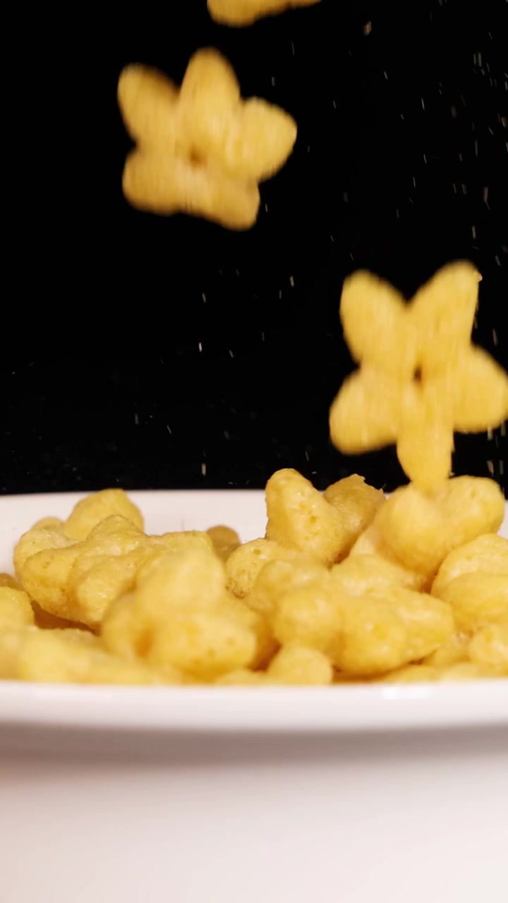 Cereais em forma de estrela caem em uma tigela branca contra um fundo preto, capturados em câmera lenta com iluminação dramática.