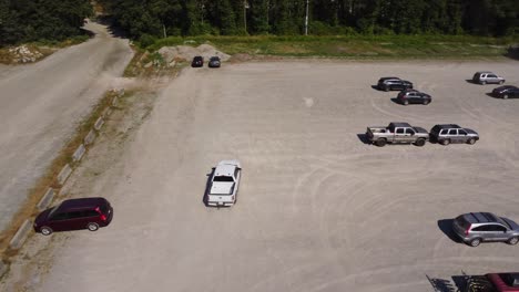 An-aerial-4K-drone-shot-follows-a-white-pick-up-truck-as-it-drifts-and-makes-donuts-in-an-event-parking-lot,-kicking-up-dust-amidst-trees-in-Squamish,-BC