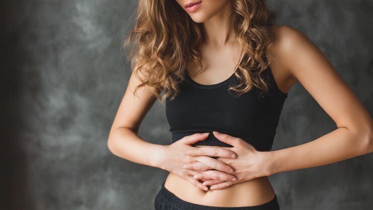 A woman with curly hair in a black top holds her stomach, visibly expressing discomfort or concern in an intimate indoor setting against a gray backdrop