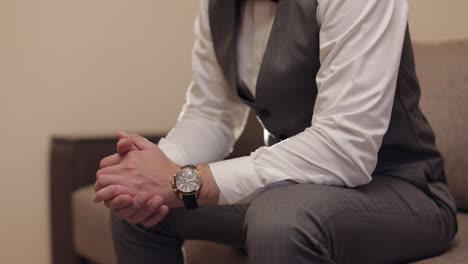 a groom in a suit and vest, sitting with his hands folded on his lap. he is wearing a watch on his wrist.