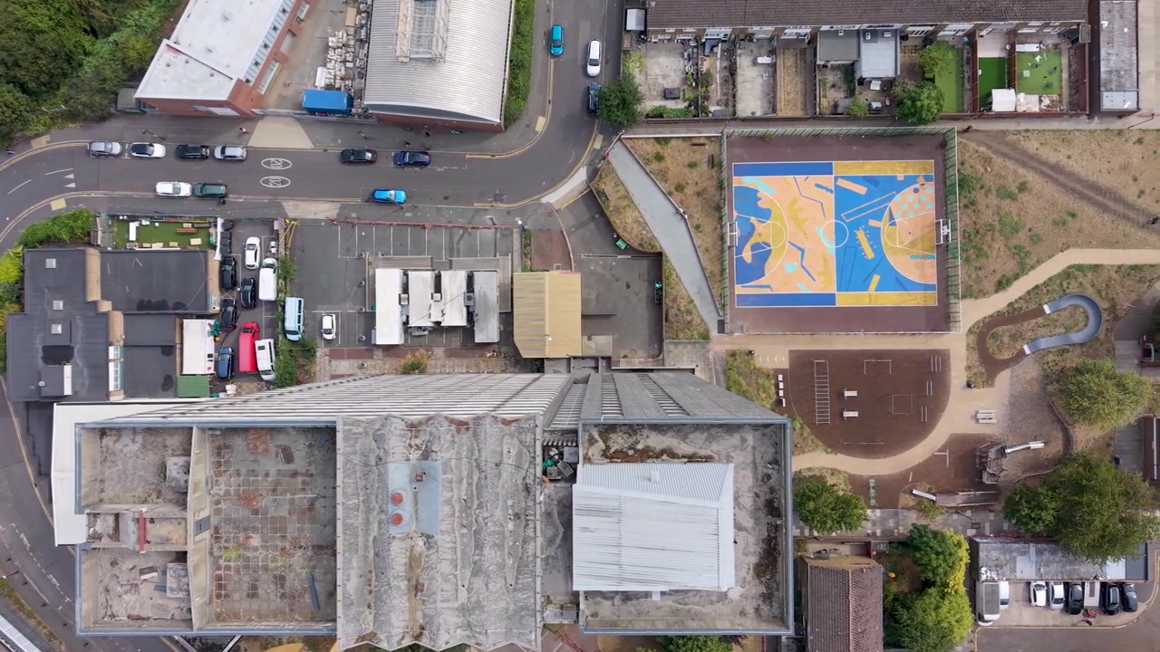 Palazzo fatiscente dall'alto mentre un drone scende in picchiata su strade del centro città, auto parcheggiate, tetti, case e un luminoso campo sportivo polivalente in un contesto di quartiere urbano