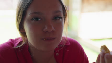 close up of young woman in red shirt savoring flaky snack with closed eyes, focused on flavor and texture, sunlight streaming from background window