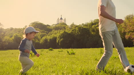 Un-Joven-Padre-Con-Una-Camiseta-Blanca-Con-Dos-Hijos-Jugando-Al-Fútbol-En-El-Césped-Al-Atardecer-Bajo-El-Sol-En-Cámara-Lenta