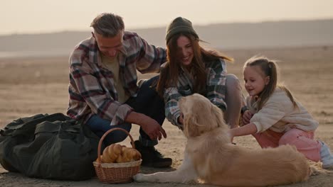 A-happy-brunette-man-with-gray-hair-in-a-checkered-shirt-along-with-his-wife-and-little-daughter-play-with-their-domestic-large-cream-colored-dog-during-their-picnic-on-a-deserted-seashore-in-the-summer