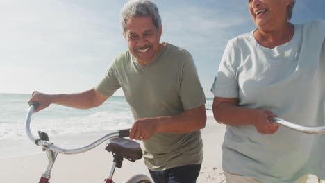 happy hispanic senior couple walking with bikes on beach at sunset