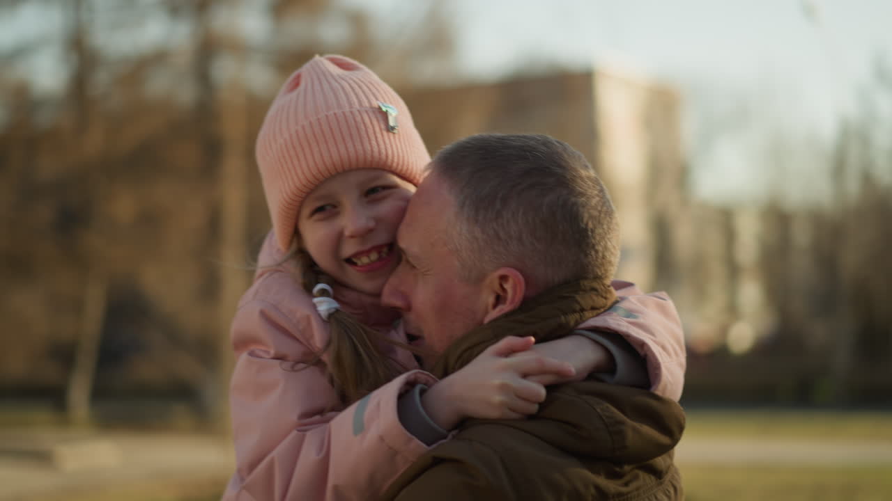 un hombre con una chaqueta marrón lleva a una niña con una gorra rosada y una chaqueta mientras ella lo abraza fuertemente. la escena captura un momento tierno y amoroso de unión padre-hija al aire libre en un día soleado