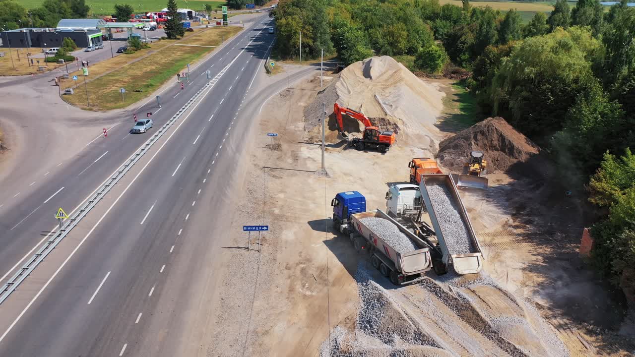 Trucks pouring out granite stone. Heavy machinery carry metal for construction new asphalt road near the highway. Aerial view.