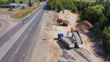 Trucks pouring out granite stone. Heavy machinery carry metal for construction new asphalt road near the highway. Aerial view.