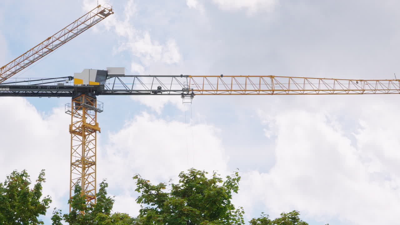 Yellow industrial tower crane operating on a construction site against a cloudy sky background. Building machinery lifting heavy loads