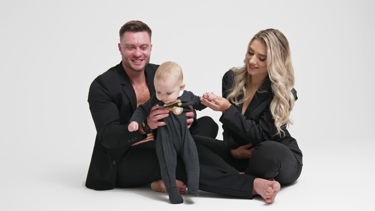 Caucasian family of three spending time together. Parents wearing dark suits sit on the floor in studio holding their adorable baby. White backdrop.