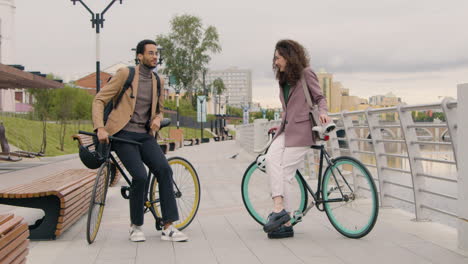 american man and woman talking to each other while leaning on their bikes on the city bridge