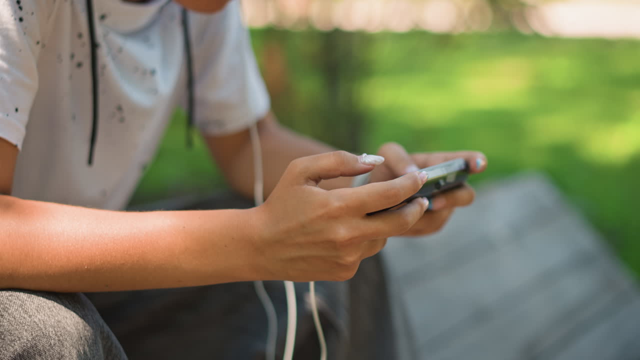 Primer plano de unas manos asiáticas sujetando el mando de un smartphone que muestra los movimientos del pulgar y el cable que lleva los auriculares; toma de acción del juego móvil, precisión y ritmo en un banco del parque, ideal para practicar streaming