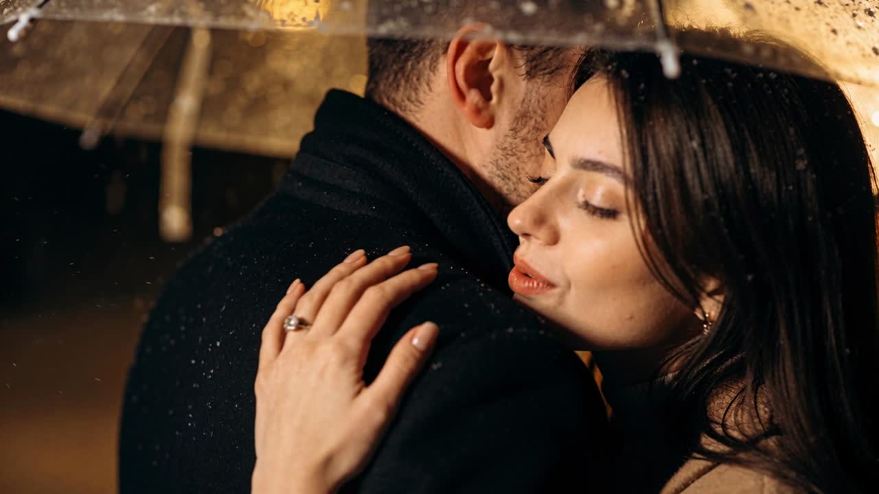 Pareja acurrucada bajo un paraguas transparente de noche, empezando a llover; el hombre con abrigo y bufanda, la mujer mostrando un anillo.