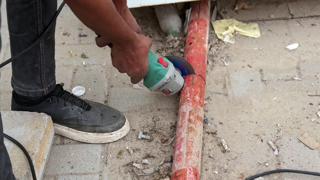 Primer plano de un trabajador usando una amoladora angular para cortar un tubo metálico oxidado en una obra polvorienta, con chispas visibles y manejo manual a plena luz del día.