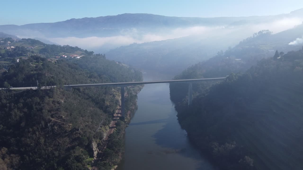 Aerial view of a long, modern bridge spanning a wide river valley, with lush green, misty mountains rising dramatically on both sides under a hazy sky.