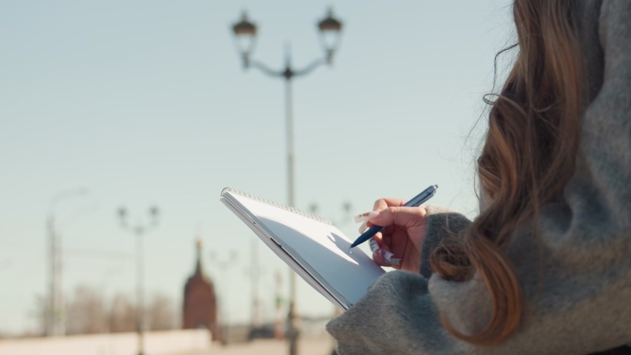 Silent outdoor journaling. Person sketches city skyline as daylight fades and night approaches. Woman records thoughts in her notebook while sitting beside river illuminated by streetlamps