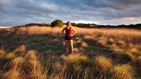 Young-woman-posing-on-the-side-of-a-hill-after-a-workout-at-sunrise-drone-shot-pull-out