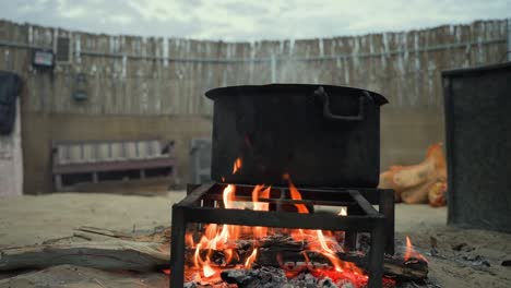 static view of a large black pot over glowing embers in a rural setting