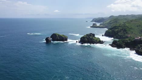 drone shot of overgrown rocks in ocean,hit by waves during sunny day - timang island, yogyakarta, indonesia