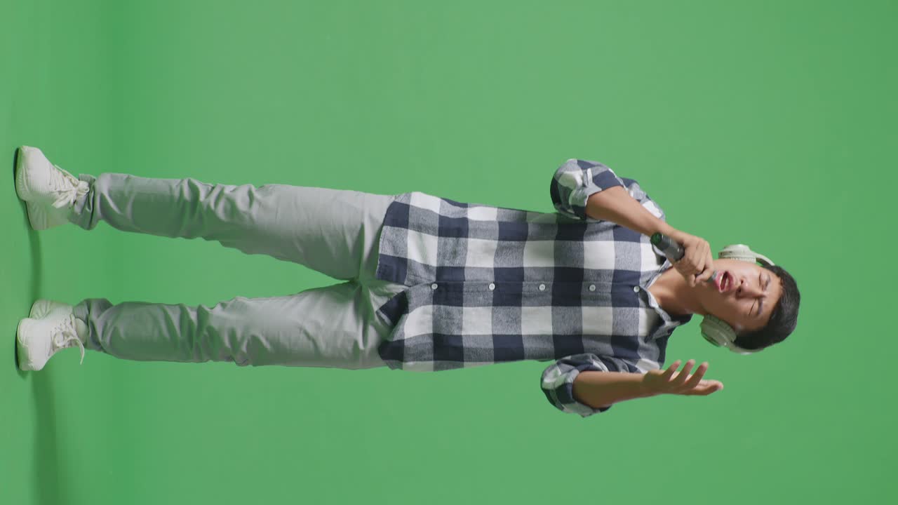 Full Body Of Young Asian Teen Boy Wearing Headphone And Holding A Microphone And Singing On The The Green Screen Background