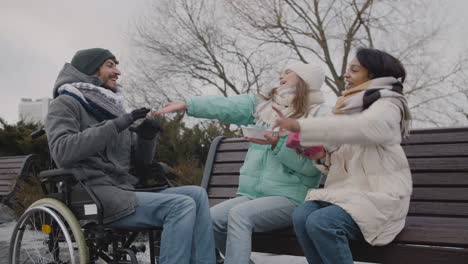 dos mujeres con un pastel pequeño y un regalo cantando feliz cumpleaños a su amigo discapacitado en el parque urbano en invierno 1