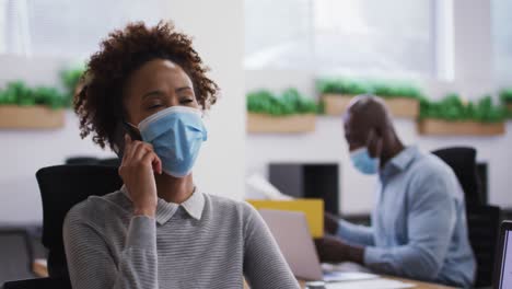 diverse male and female business colleagues in face masks, woman talking on smartphone in office