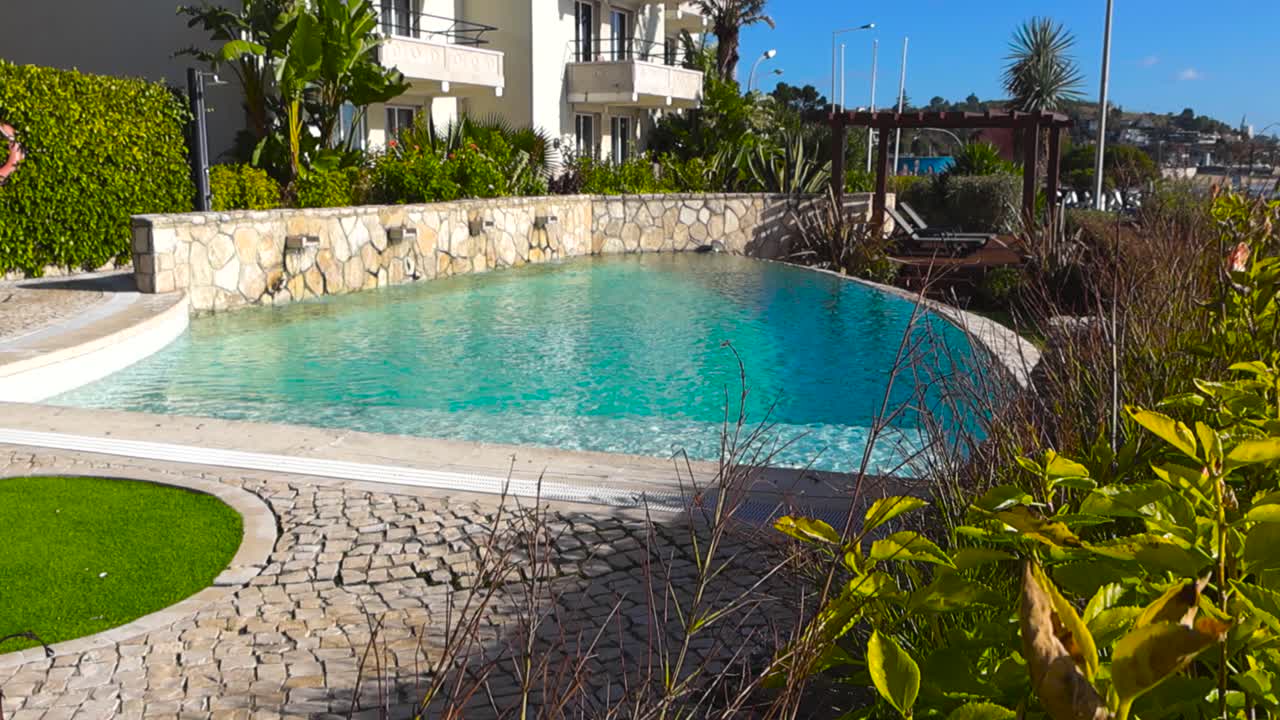 Filmatisk afsløring fra frodigt løv afslører en elegant hotelpool med turkis vand, stenbelægning og en fredfyldt flodbred på en solskinsdag