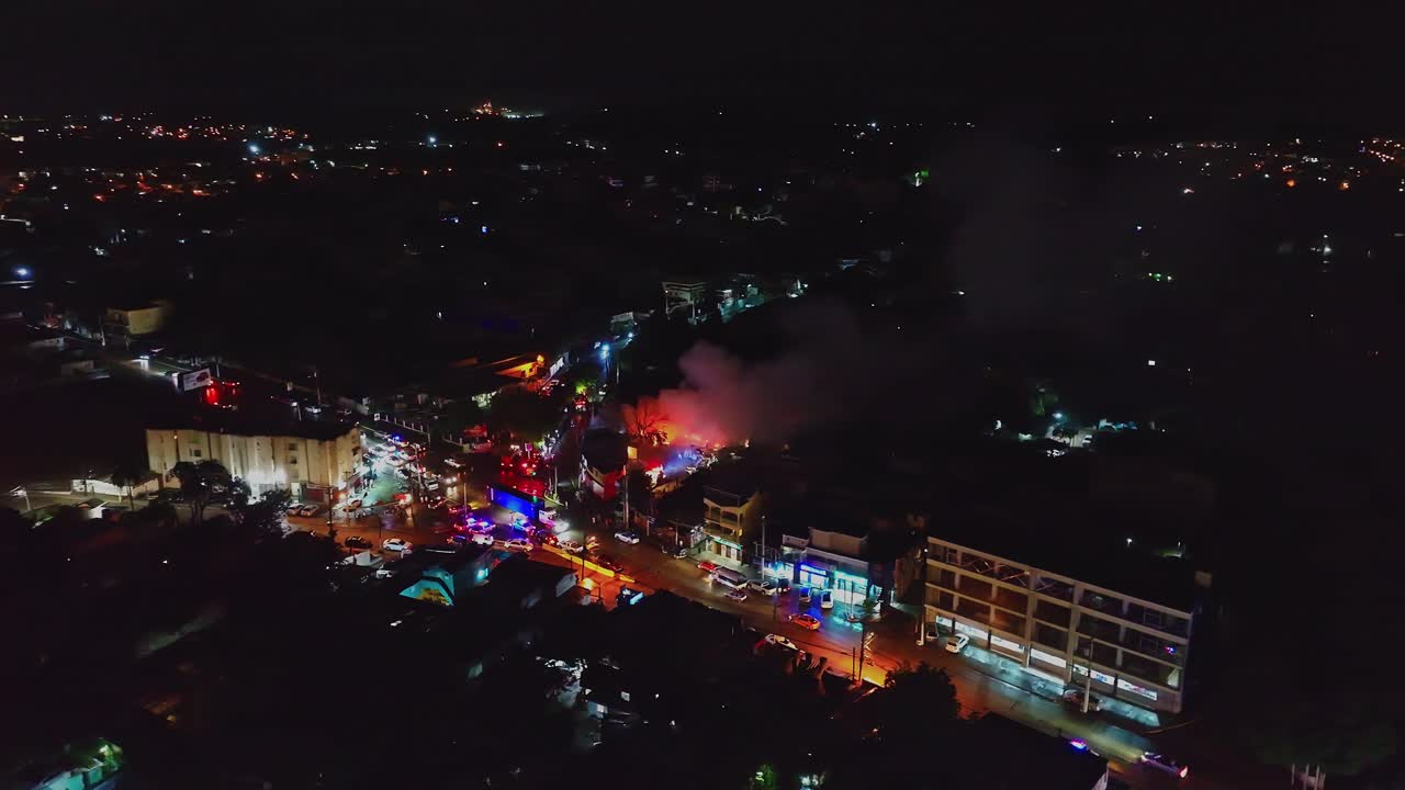 Night aerial view of warehouse fire in Santiago de los Caballeros, Dominican Republic, with flames, heavy smoke and emergency vehicles responding in illuminated urban area. Top down shot