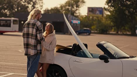 A-blond-guy-with-glasses-in-a-plaid-shirt-stands-near-his-blonde-girlfriend-in-a-white-sweatshirt-and-kisses-her,-holding-hands-near-a-white-convertible-with-a-surfboard