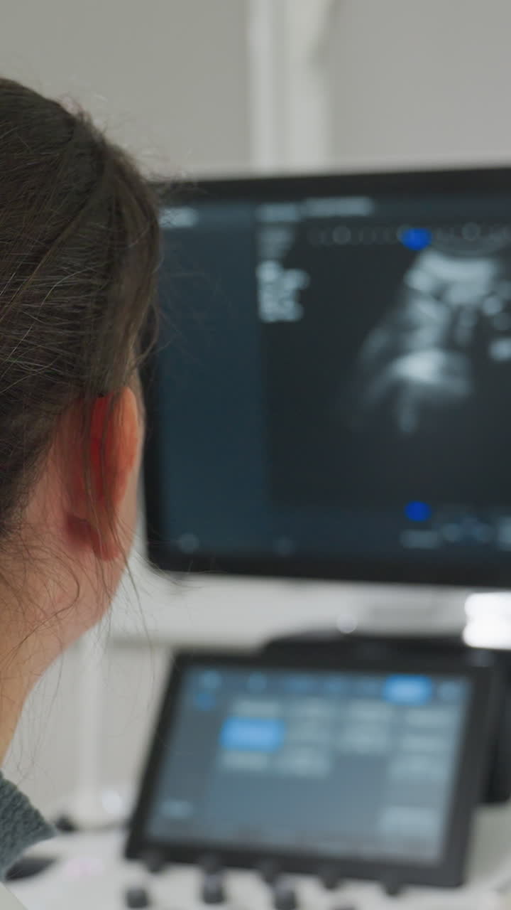 black woman watching ultrasound monitor screen in clinic with technician operating console, grayscale fetal image visible on display, touchscreen interface in foreground, rear profile of female