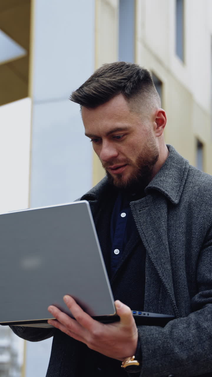 Businessman with a laptop outdoors. Handsome man in warm coat standing in the city street and working on a laptop. Business and work concept. Vertical video
