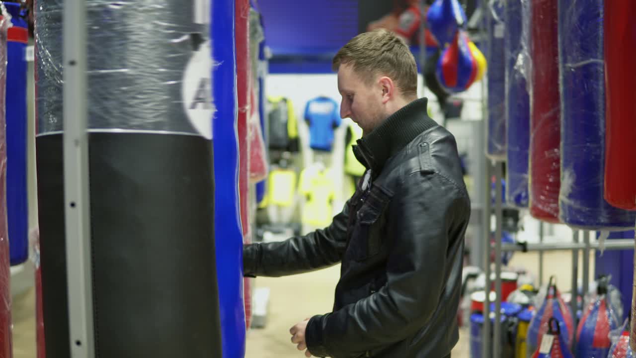 un hombre adulto con chaqueta de cuero negro eligiendo un saco de boxeo en una tienda de deportes