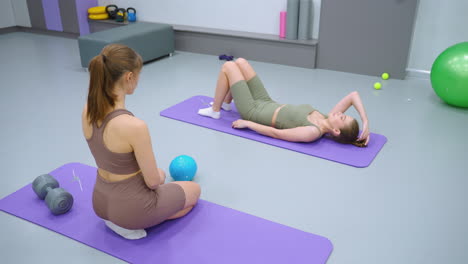 mujer acostada en una alfombra púrpura ajustando su cabello mientras su amiga se arrodilla cerca, observando, globos de colores llenan el fondo, añadiendo un toque lúdico a esta escena de fitness o relajación