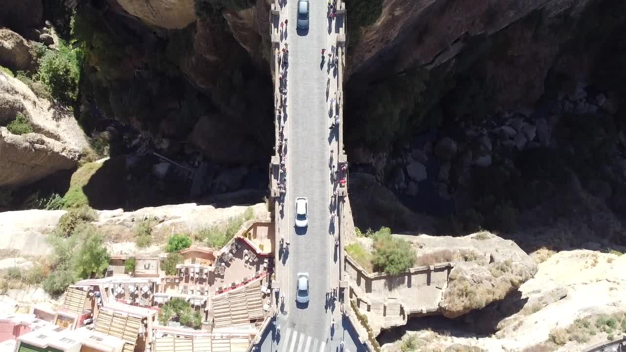 Top down drone view of Puente Nuevo in Ronda shows narrow bridge with pedestrian and vehicle movement spanning deep rocky gorge surrounded by cliffs and buildings on both ends under bright daylight