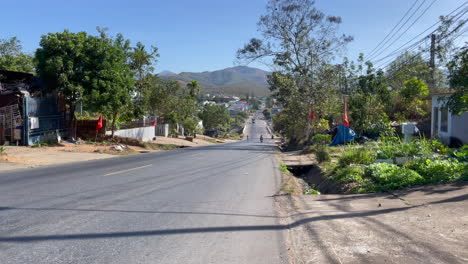 People-riding-mopeds-through-village-in-Vietnam-with-mountains-in-background,-Kom-Yum-Province