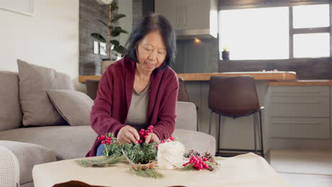senior asian woman crafting christmas wreath at home, focusing on holiday decorations