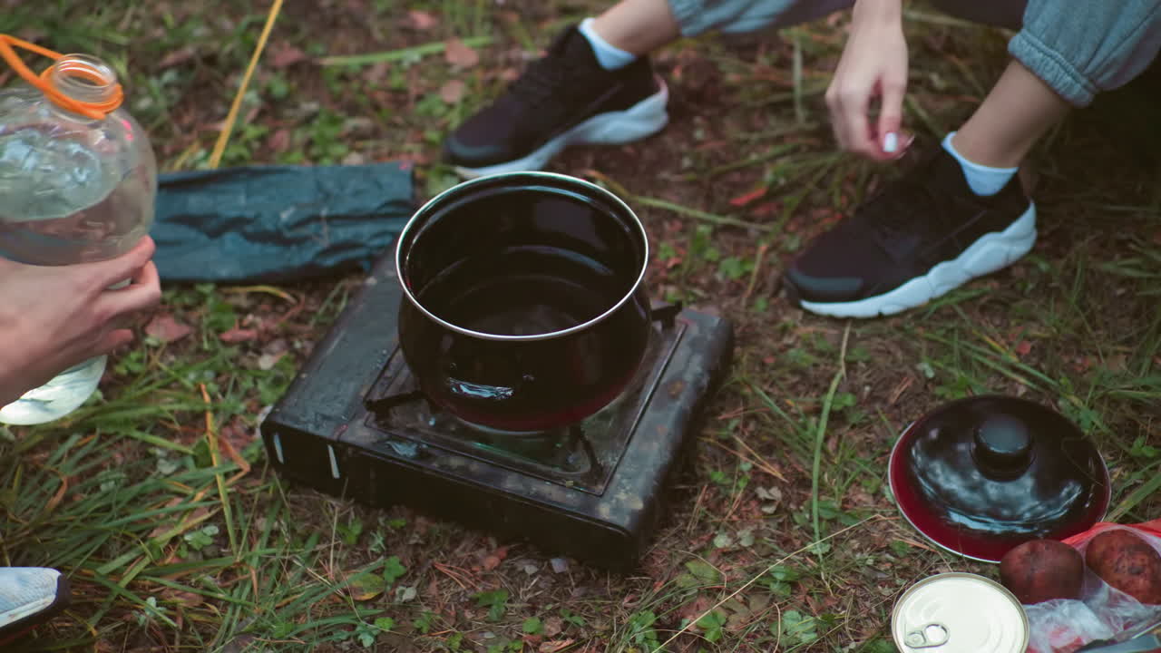 overhead view of campers cooking in forest as woman lights portable gas stove and another person pours water into black cooking pot beside scattered food items and camping gear on grass
