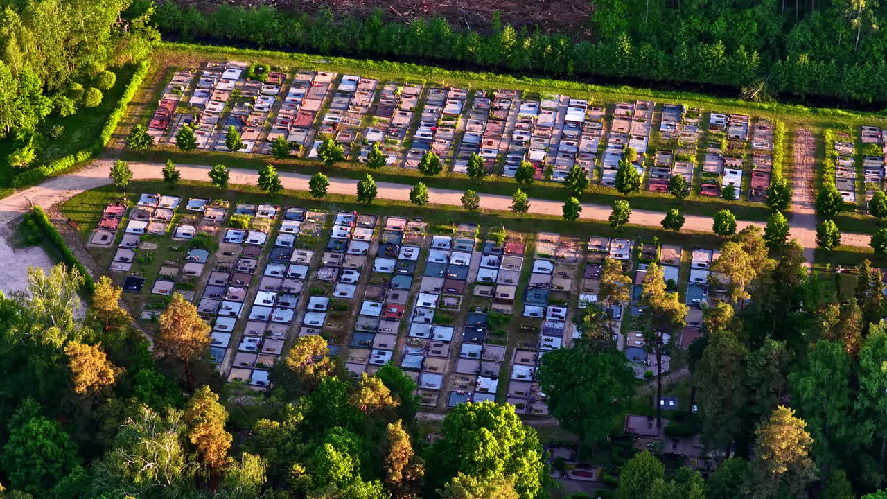 Aerial drone view of a peaceful cemetery surrounded by dense green forest. Rows of graves and pathways create a structured pattern, captured in warm evening light from above