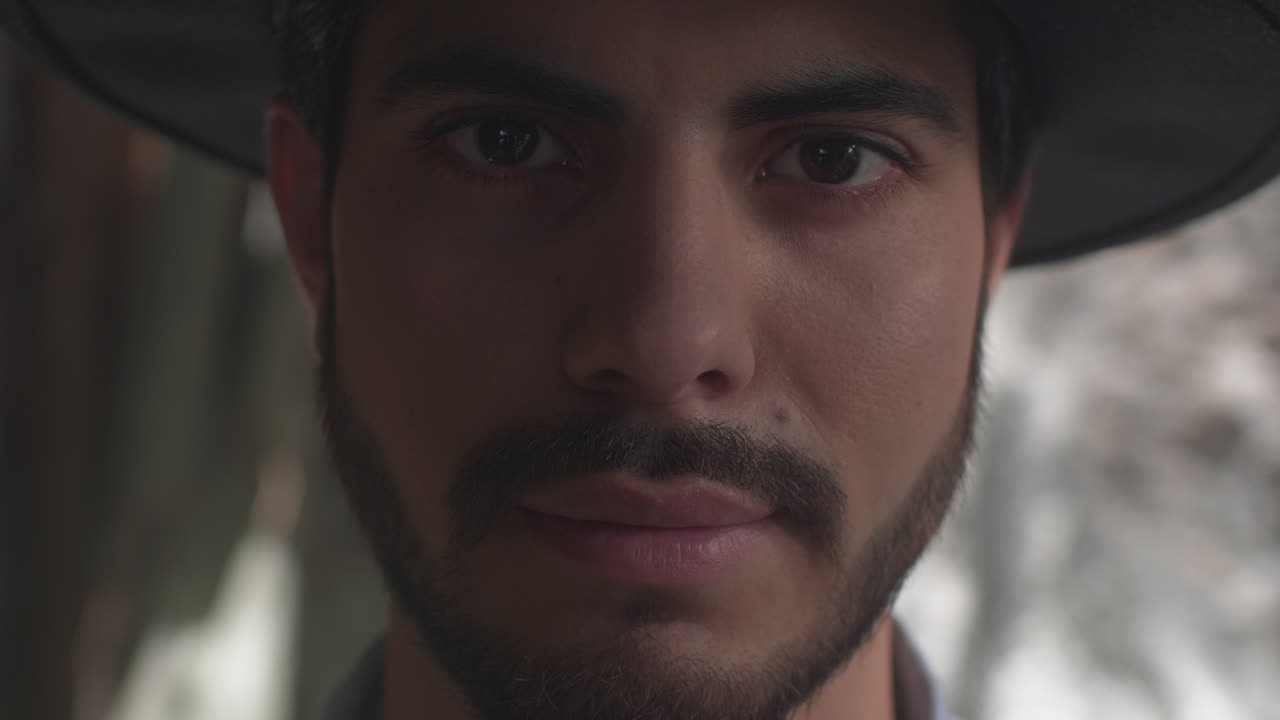 High-quality 4K footage of a stylish young man with a beard and hat, slowly opening his eyes in soft, natural light. He has a focused, mysterious expression. Ideal for travel, discovery