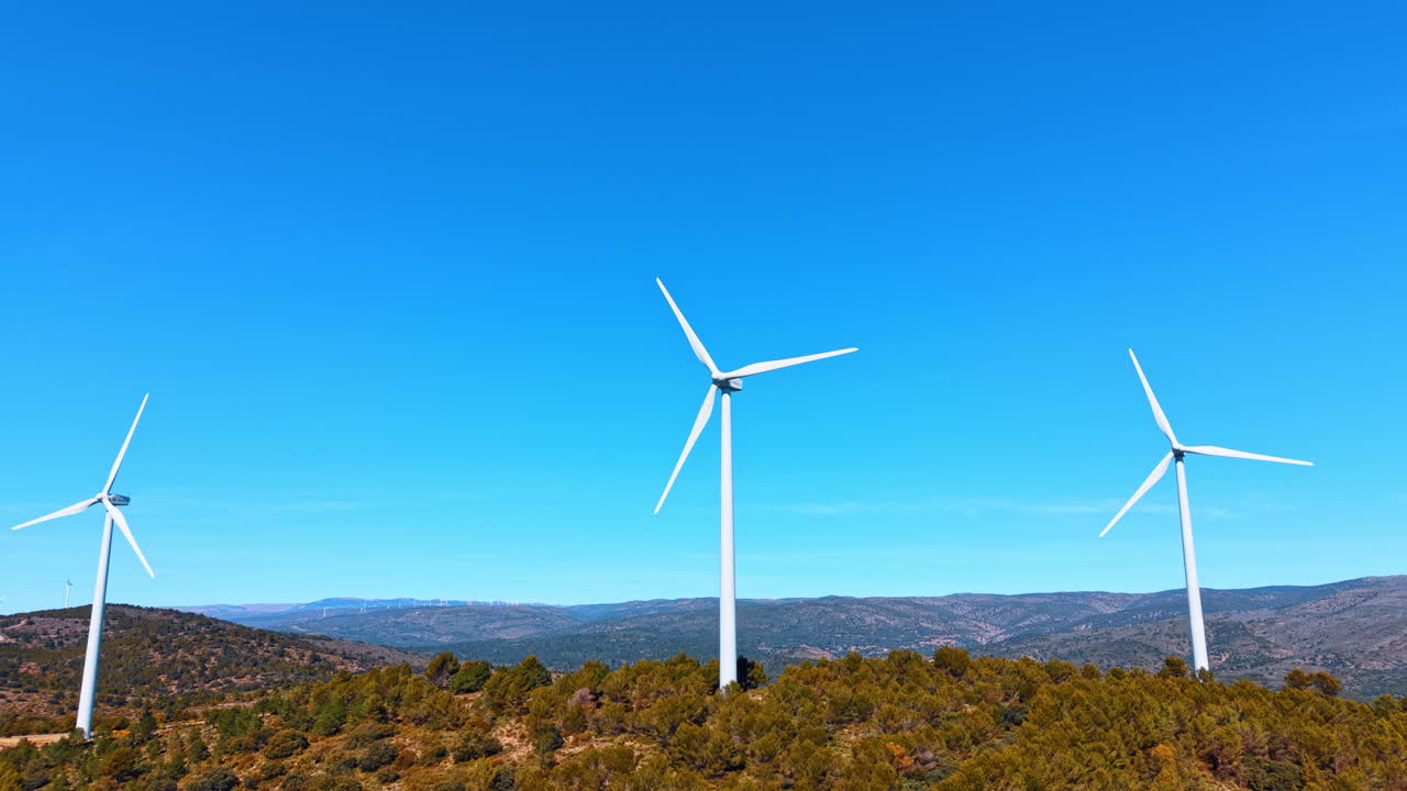 Madrid, Spagna, 8 agosto 2025: Tre aerogeneratori bianchi su colline boscose sotto un cielo blu. Ripresa aerea di un parco eolico su crinali verdi, orizzonte limpido, infrastruttura per l'energia rinnovabile, piena luce diurna.