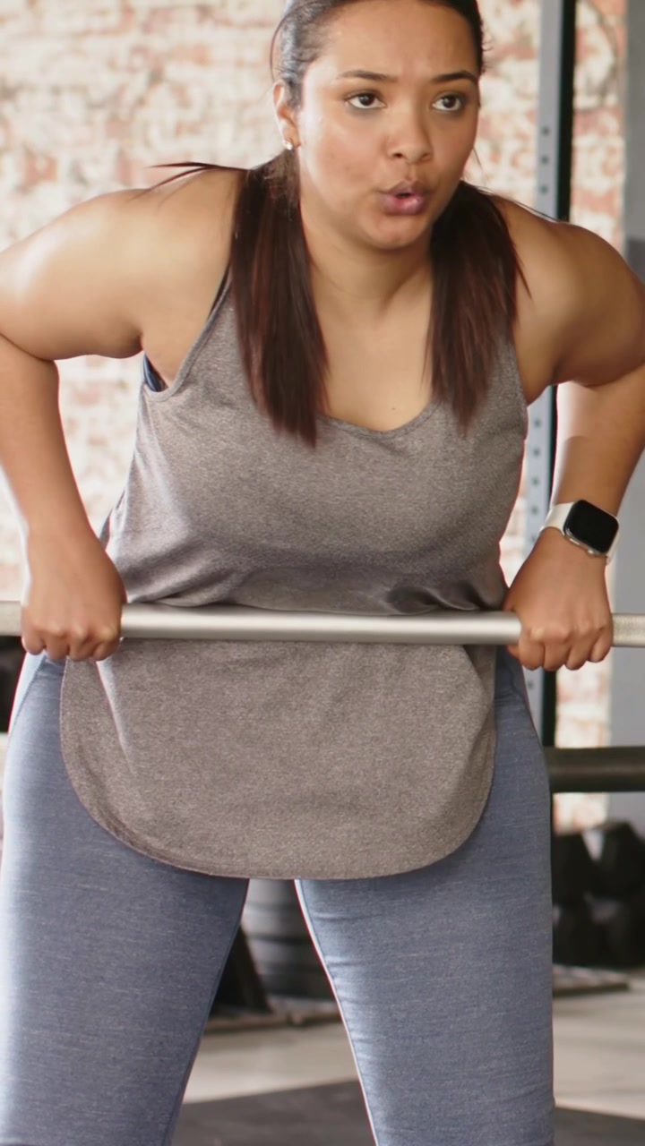 Video verticale: Donna in canotta grigia che afferra un bilanciere ed esegue rematore a busto flesso in palestra per la forza.