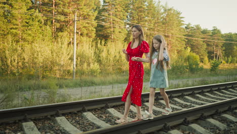 side view of woman holding hand of young girl during peaceful evening walk on railway track surrounded by lush green forest trees and power lines under warm golden sunlight in countryside environment