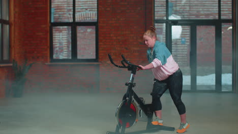 Chica-Atlética-Realizando-Ejercicios-De-Entrenamiento-Aeróbico-En-Bicicleta-Estática-En-Un-Gimnasio-Con-Niebla