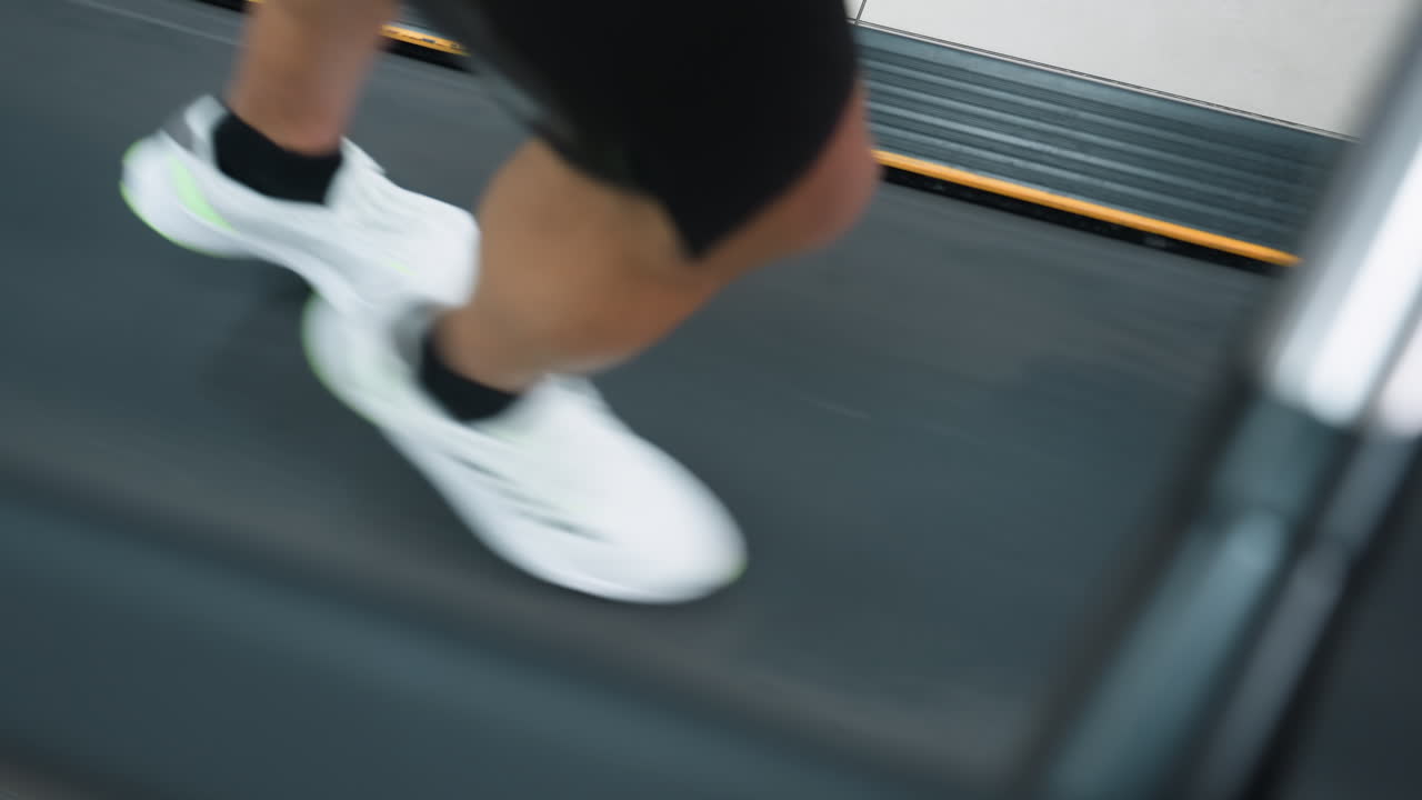 top down view of man running on treadmill belt, motion blur captures white sneakers on textured surface, gym equipment edges visible, window light reflection adds contrast to fitness scene